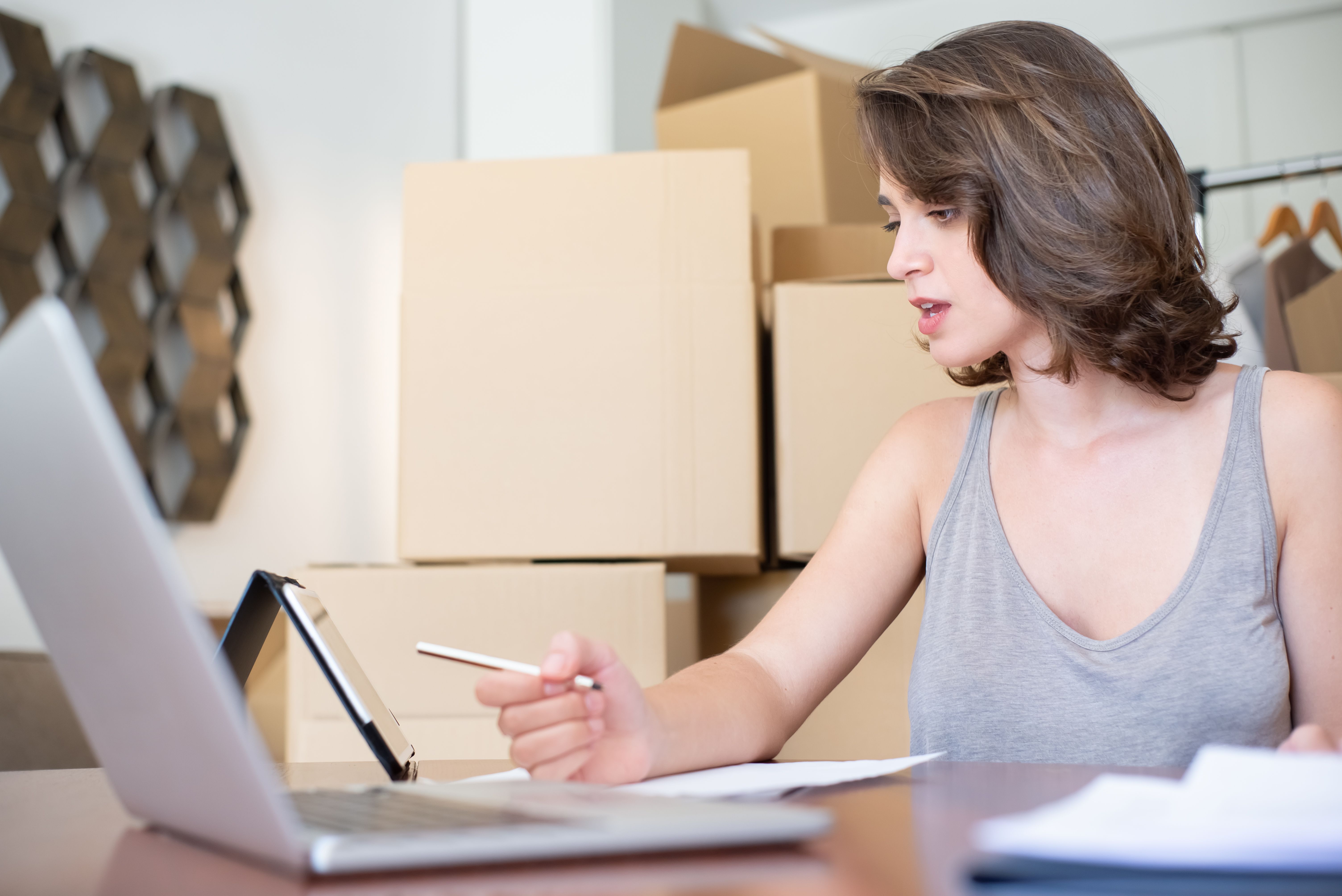 Woman managing online store orders with a laptop and tablet among shipping boxes.