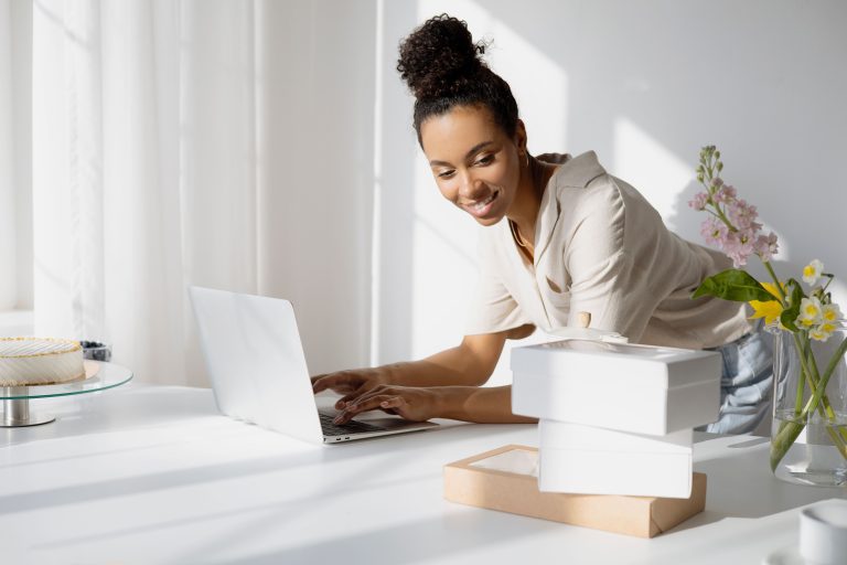 Woman entrepreneur managing an online shop from a bright home office with product boxes and flowers.