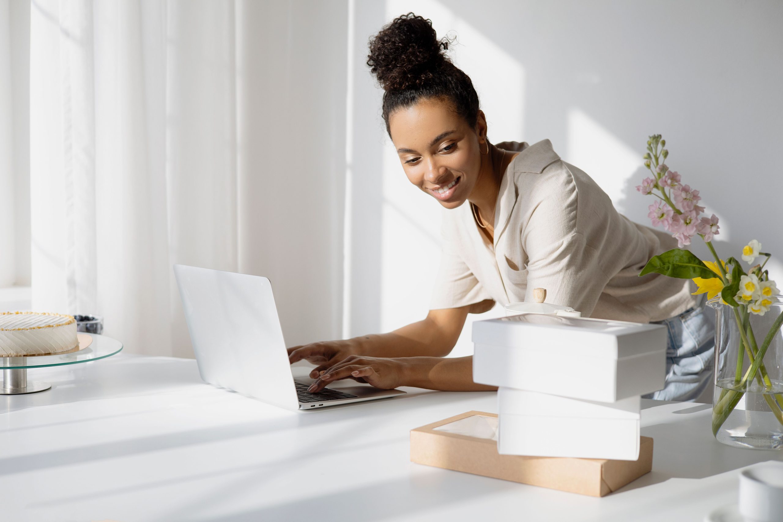 Woman entrepreneur managing an online shop from a bright home office with product boxes and flowers.